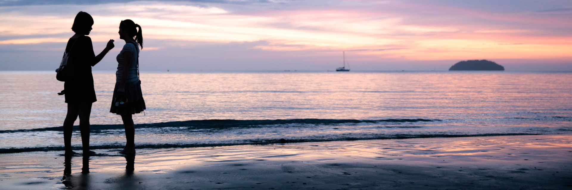 Deux femmes discutent sur un fond de couché de soleil au bord de la plage. Le ciel est mauve et chaud. Une ile est visible dans le second plan avec un bateau. 