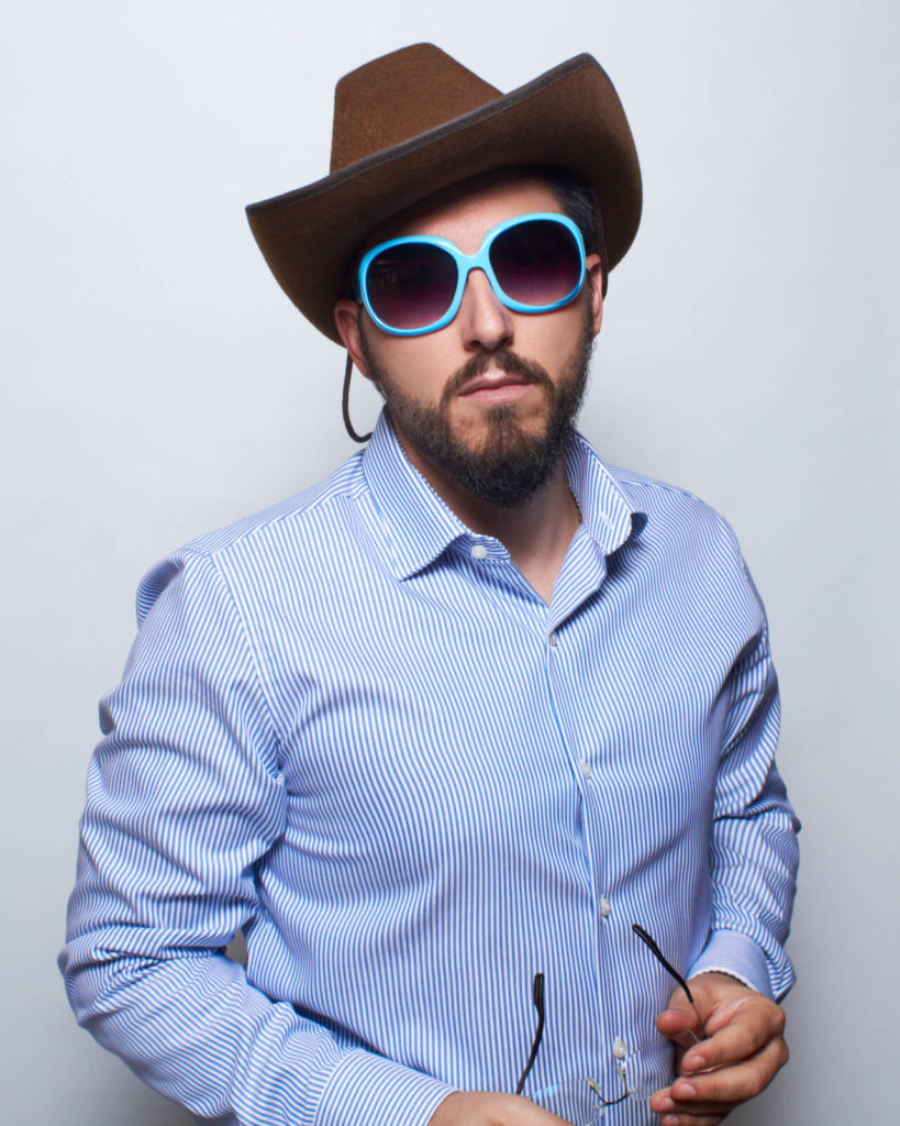 Un homme avec une barbe porte un chapeau de cowboy marron et de grandes lunettes de soleil bleues. Il est devant un fond simple, habillé d'une chemise à rayures. Il tient les lunettes normales dans une main et regarde directement la caméra avec une expression neutre.