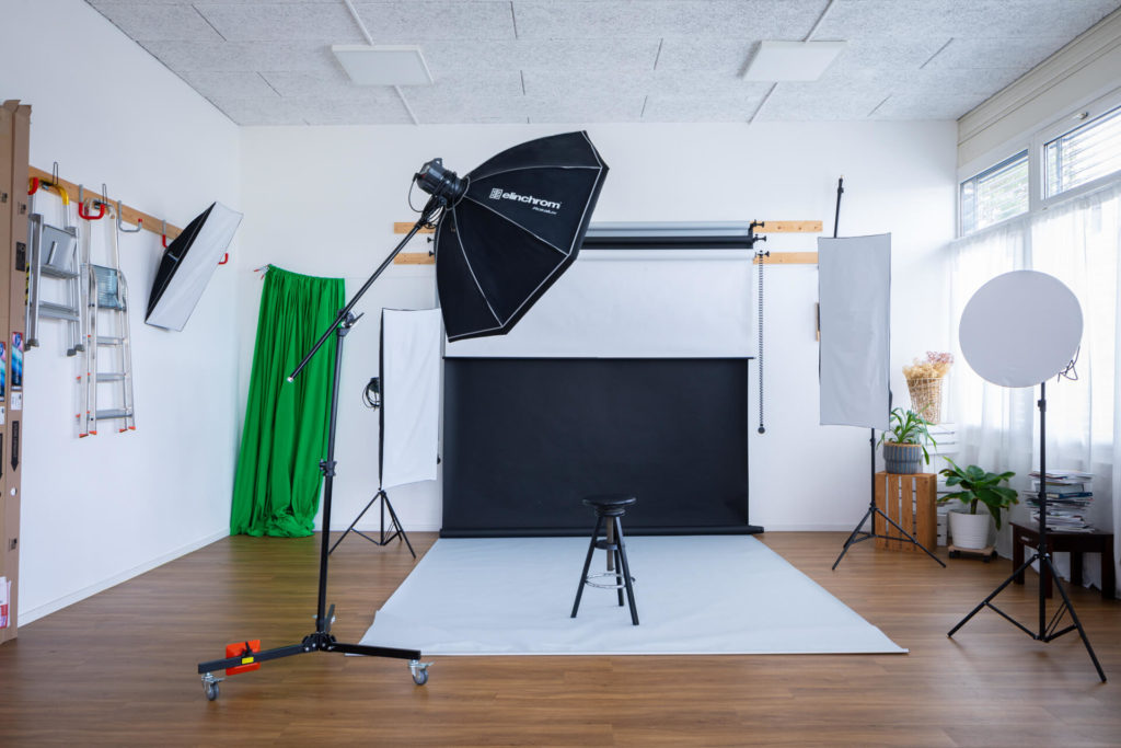 Le studio a divers équipements d'éclairage comme des boîtes à lumière, des parapluies et des réflecteurs. La pièce a un sol en bois et des murs blancs, avec de la lumière naturelle qui entre par une fenêtre.