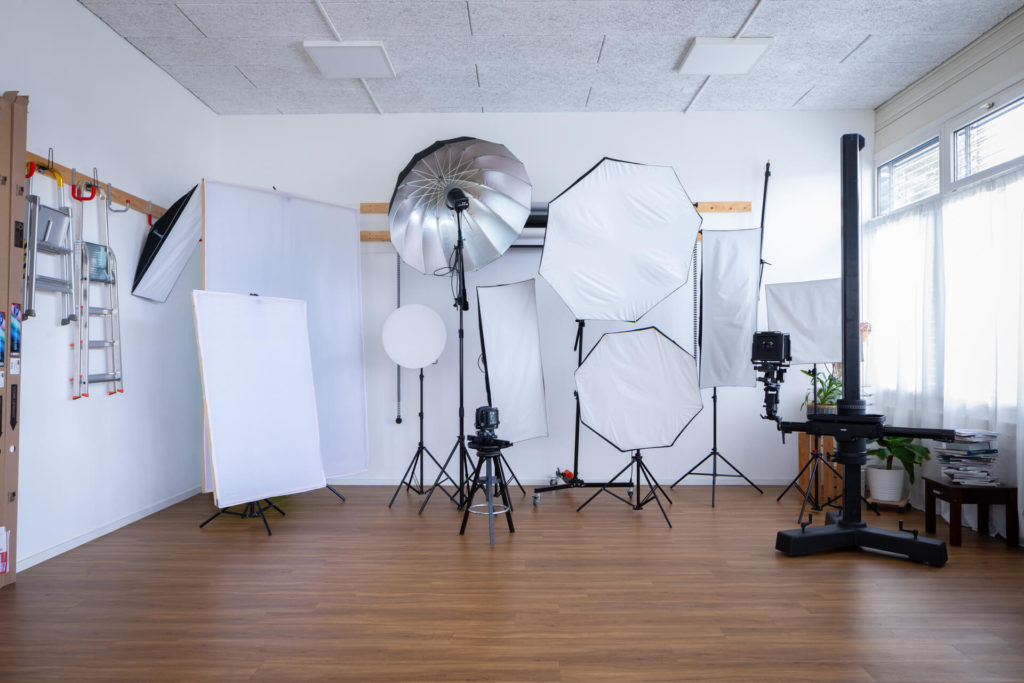 Le studio a divers équipements d'éclairage comme des boîtes à lumière, des parapluies et des réflecteurs. La pièce a un sol en bois et des murs blancs, avec de la lumière naturelle qui entre par une fenêtre.