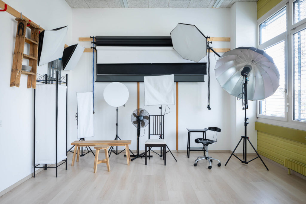 Le studio a divers équipements d'éclairage comme des boîtes à lumière, des parapluies et des réflecteurs. La pièce a un sol en bois et des murs blancs, avec de la lumière naturelle qui entre par une fenêtre.