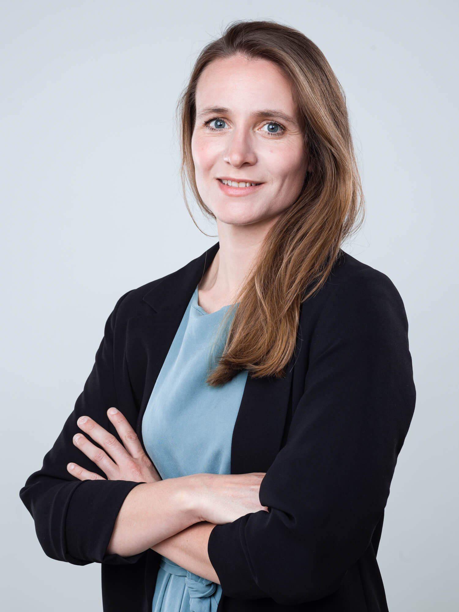 Une femme aux longs cheveux bruns est debout et sourit doucement, vêtue d'un blazer noir sur un chemisier bleu sarcelle sur un fond gris clair. Elle a croisé les bras dans son portrait professionnel.