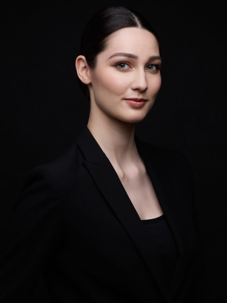 Une femme aux cheveux lisses et au maquillage minimal est photographiée par des professionnels. Elle est vêtue d'un blazer noir, debout sur un fond sombre, souriant légèrement alors qu'elle regarde directement la caméra.