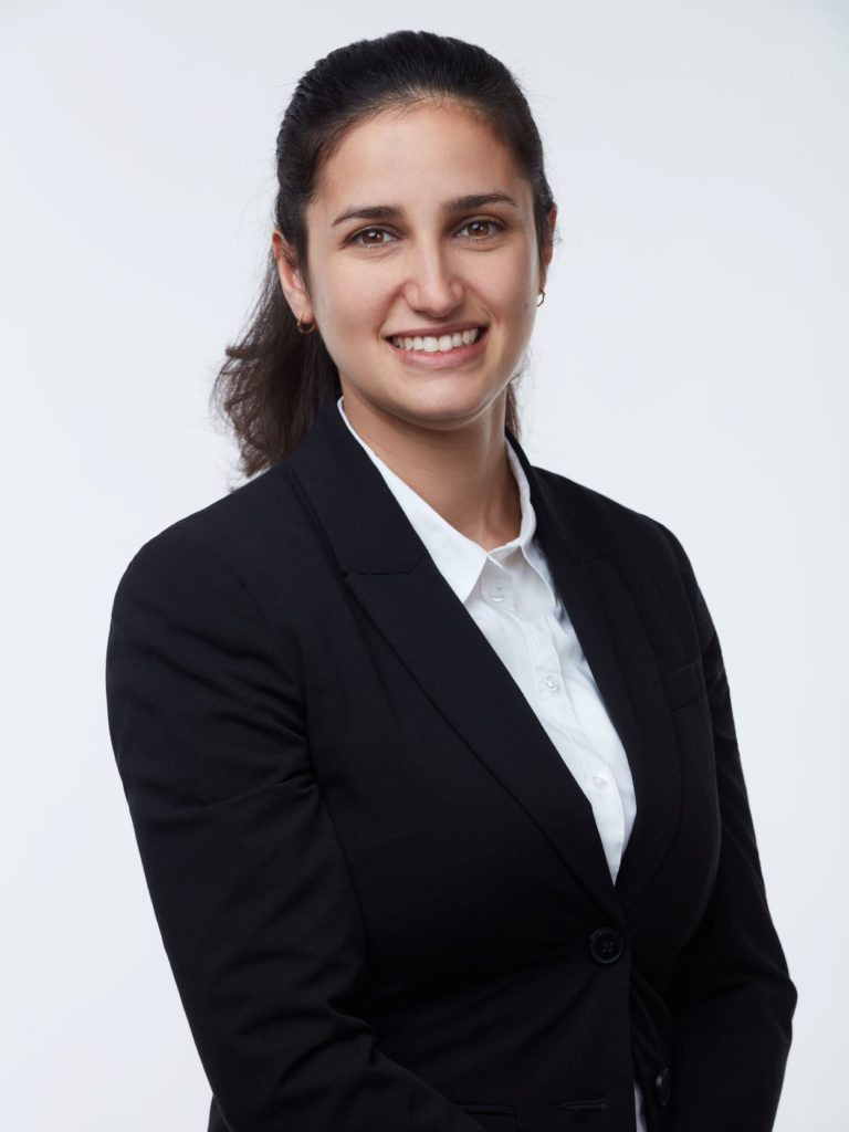 Une femme souriante aux longs cheveux noirs est photographiée professionnellement. Elle porte un costume noir et une chemise blanche sur fond gris clair.