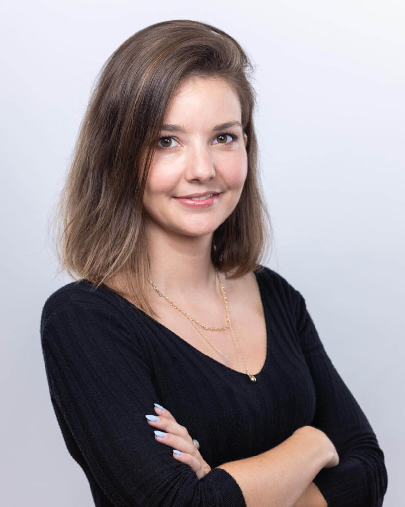 Une photo professionnelle montre une femme souriante aux cheveux bruns mi-longs. Elle porte un haut noir et un collier fin et se tient devant un fond gris clair.